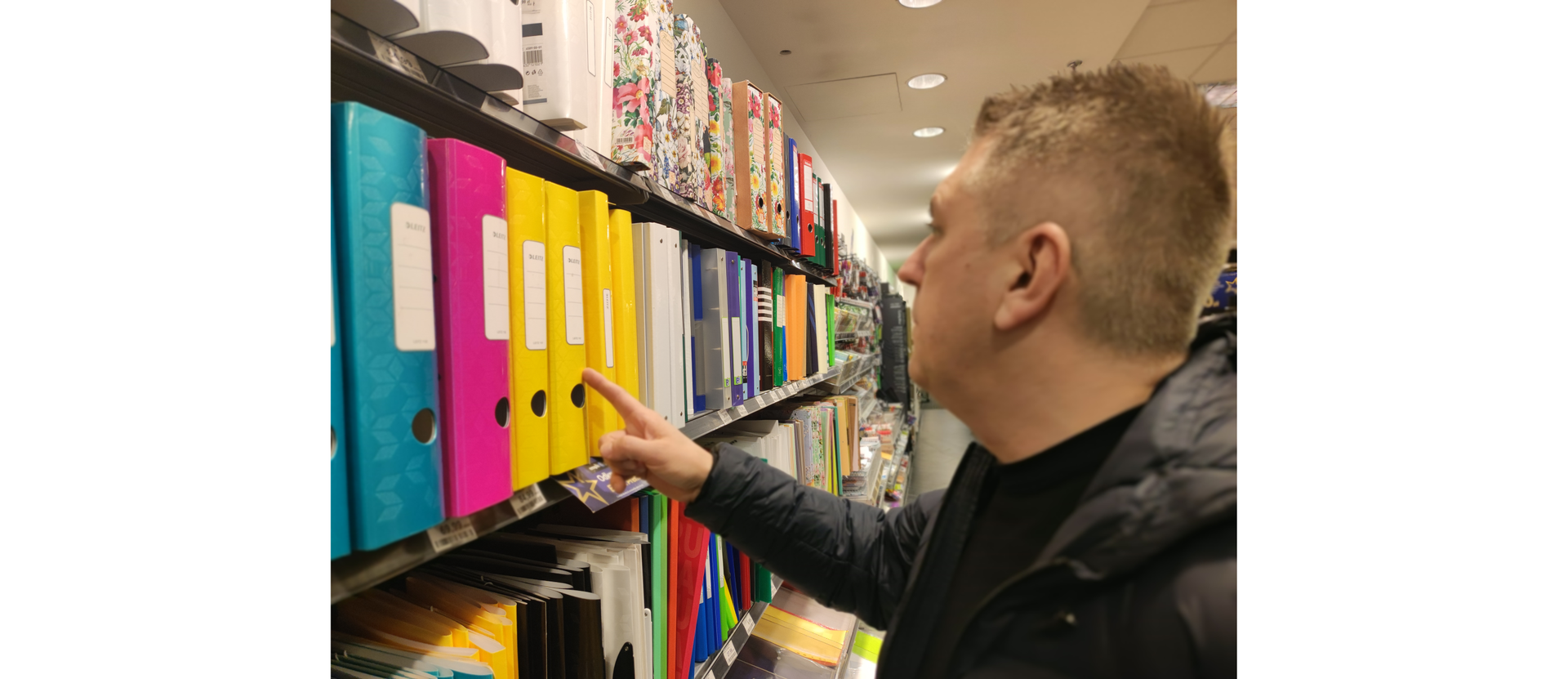 Zdjęcie przedstawia mężczyznę w sklepie papierniczym oglądającego segregatory na półce.