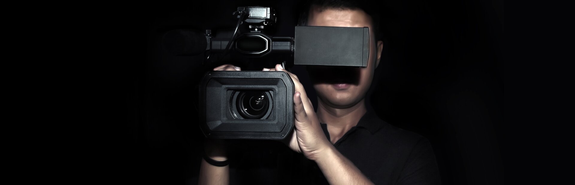 Fotografia przedstawia mężczyznę trzymającego kamerę na prawym ramieniu. Jest ubrany w czarną koszulę, której górny guzik jest rozpięty. Wizjer kamery przysłania jego oczy, rzucając cień na nos oraz górną wargę. Na czole mężczyzny widać delikatne zmarszczki. Jego czarne włosy są krótkie. Kamerzysta za pomocą obu dłoni trzyma korpus kamery skierowanej do przodu. Tło jest czarne.  