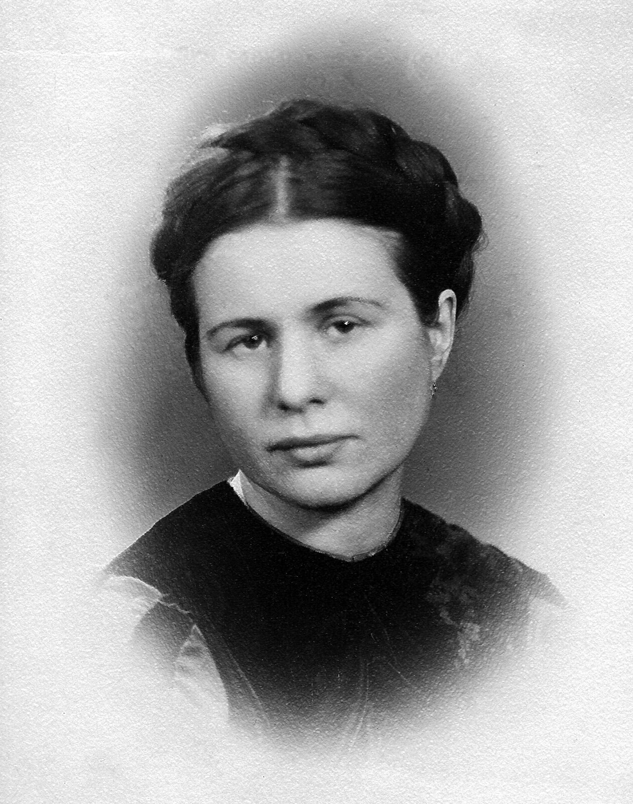  Czarno‑biała fotografia przedstawia portret młodej kobiety. Ma poważny wyraz twarzy, warkocz opleciony wokół głowy i bluzkę z dużym, ciemnym kołnierzem.