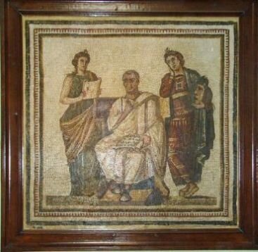 Fotografia przedstawia mozaikę oprawioną w drewnianą brązową ramę. W centrum znajduje się postać mężczyzny w długiej, jasnej szacie. Mężczyzna siedzi. Na kolanach przytrzymuje jedną ręką zwój papirusu. Po obu jego stronach stoją młode kobiety. Jedna, w czerwonej szacie i zielonym płaszczu, trzyma maskę, druga natomiast, ubrana w zieloną suknię, trzyma zwój papirusowy. Tło mozaiki jest jasne.