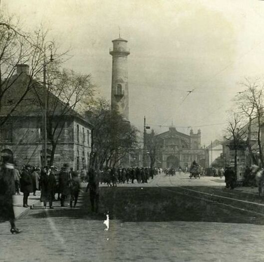 Zdjęcie przedstawia szeroką ulicę, po której biegną tory tramwajowe. Po chodnikach, wzdłuż ulicy, idą przechodnie. Na drugim planie znajduje się wieża o walcowatym kształcie.