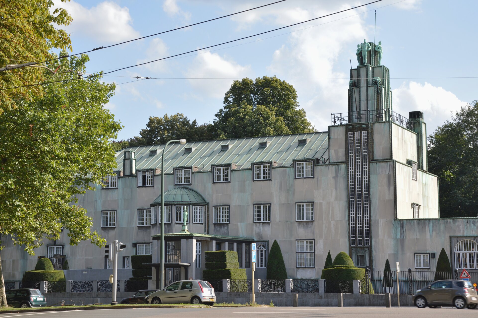 Ilustracja przedstawia budynek secesyjny Palais Stoclet autorstwa Henry'ego van de Velde. Budynek z prawej strony wieńczy wysoką wieża. Wejście do budynku mieści się na środku. Przed budynkiem i w jego tle widać zielone drzewa.