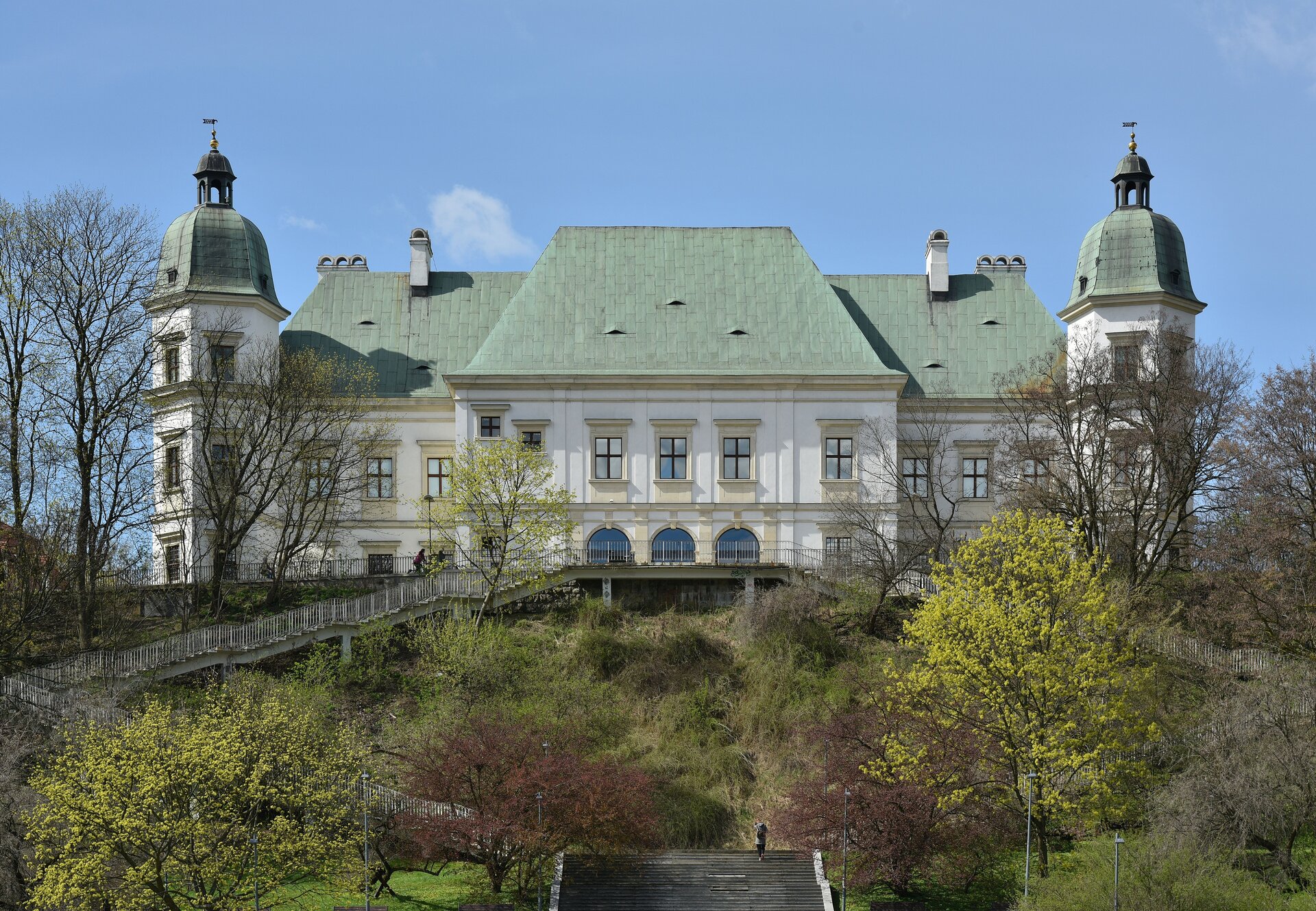 Ilustracja przedstawia Centrum Nauki Współczesnej „Zamek Ujazdowski”, w Warszawie. Na zdjęciu ukazana jest dwukondygnacyjna bryła budowli w stylu klasycystycznym z wysokim, zielonym dachem. Po obu stronach bryły znajdują się wieże przykryte kopułami, które wieńczą latarnie. Budowla ustawiona jest w scenerii zielonego parku.