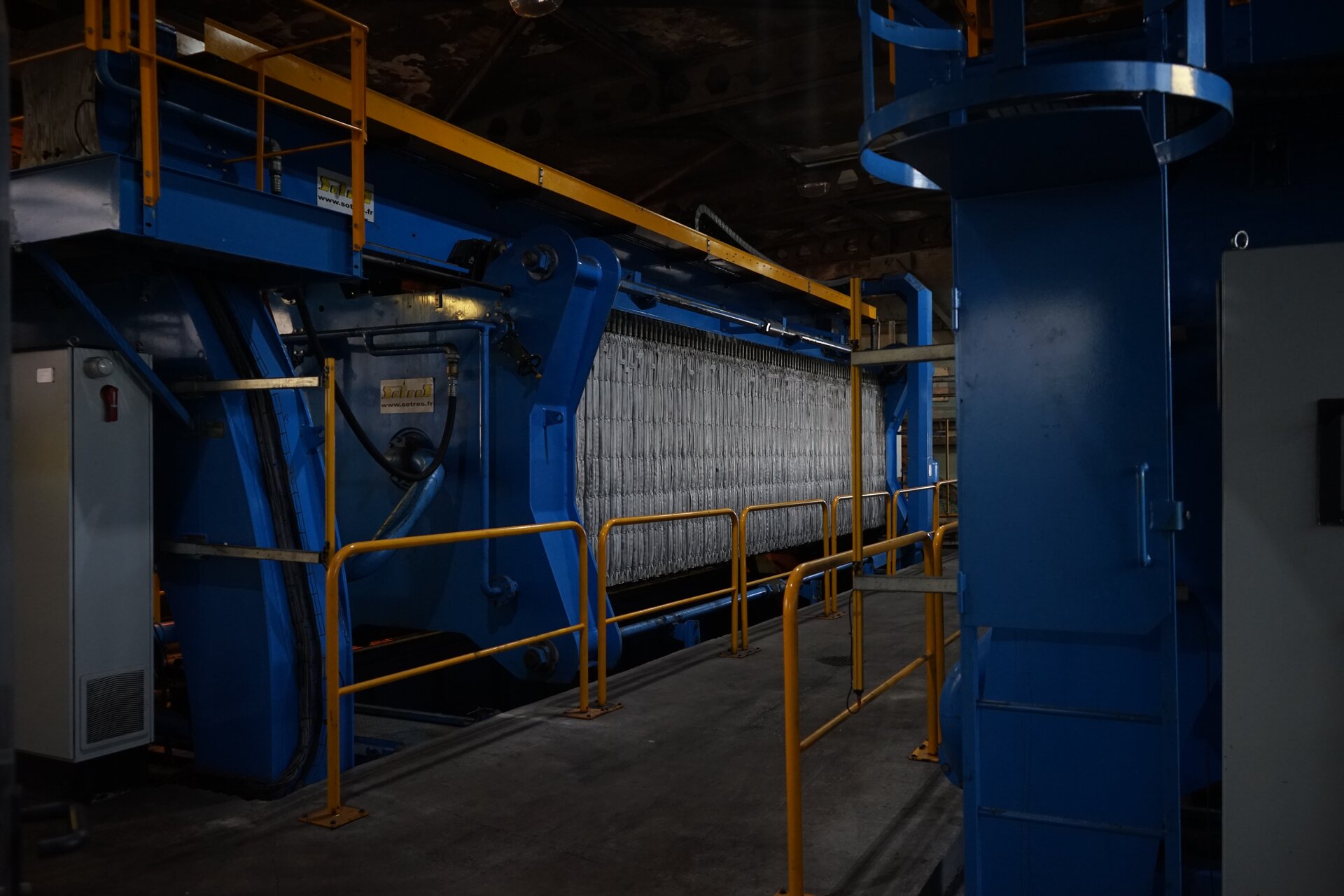 Zdjęcie przedstawia prasę filtracyjną. Jest ona wsparta na ramie. Na całej długości prostopadłościennej konstrukcji zawieszone są prostokątne przegrody. To płyty, z których wydobywane są placki filtracyjne. Prasa znajduje się po lewej stronie. Przed nią na całej długości poprowadzona jest barierka. Poniżej zdjęcia znajduje się przycisk multimedialny z nagraniem audio. Treść nagrania jest zgodna z tytułem zdjęcia.