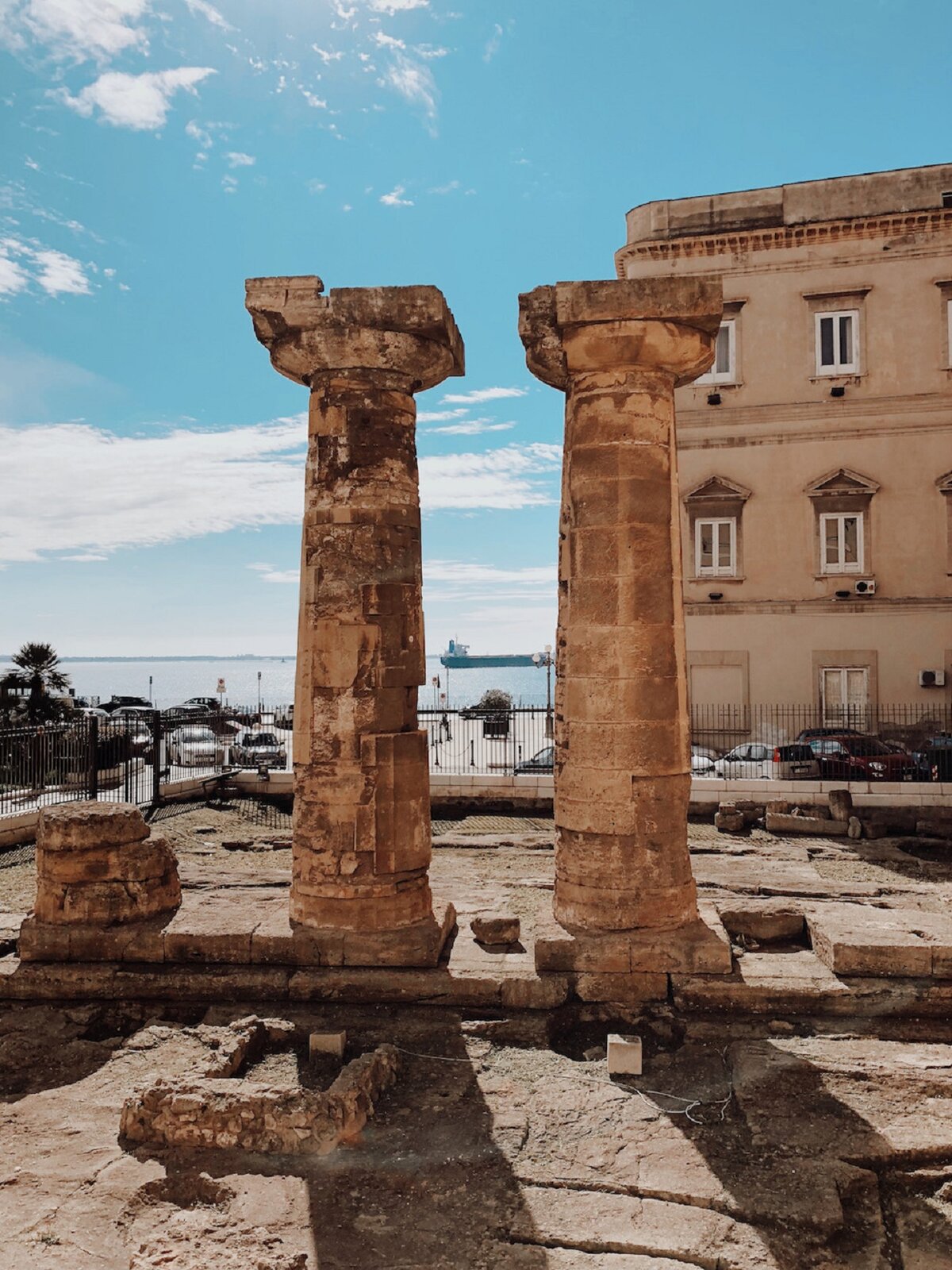 Ilustracja przedstawia fotografię „Rzymskie ruiny w Tarentum”, której autorem jest Manuela Vitulli. Na zdjęciu znajdują się beżowe ruiny z dwiema kolumnami. W tle, z prawej strony znajduje się beżowy budynek z białymi oknami, zaparkowane samochody.  W tle, z lewe strony jest morze i błękitne niebo z białymi chmurami. 