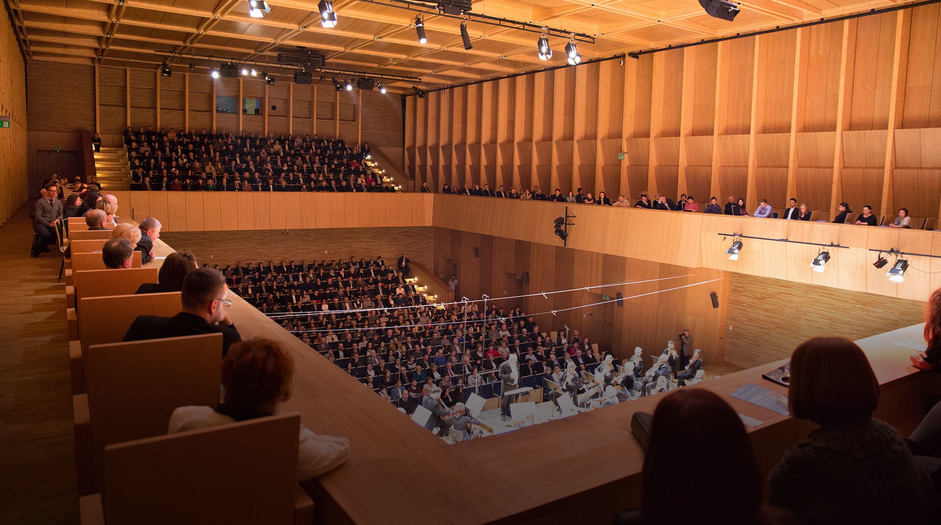 Fotografia przedstawia salę koncertową, która ukazana jest z perspektywy widza siedzącego na balkonie okalającym całą salę. W dole scena z grającą orkiestrą, w tle publiczność. Cała sala utrzymana jest w kolorze jasnego drewna.