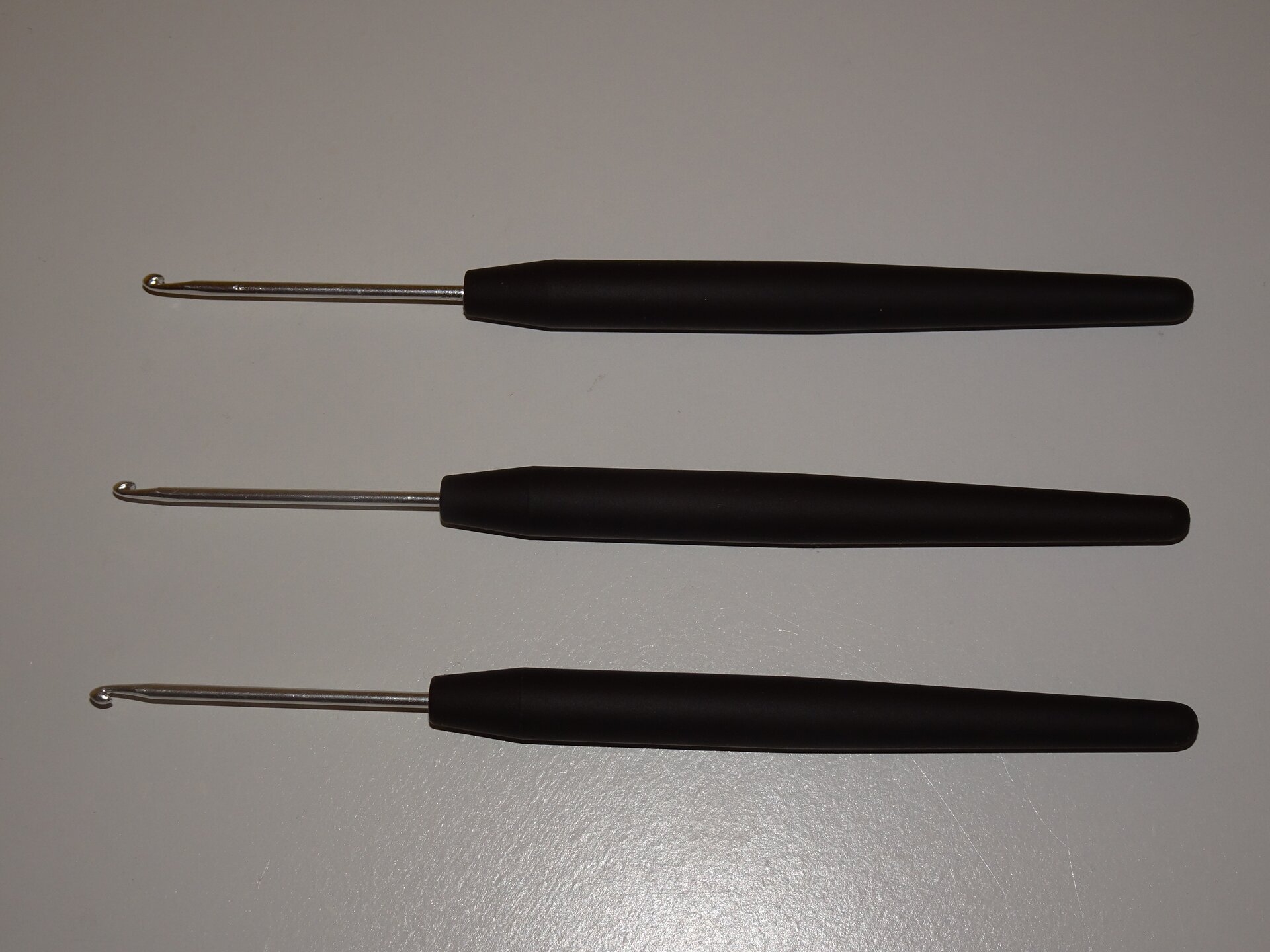 Zdjęcie przedstawia trzy szydełka. Każde szydełko składa sie z metalowego pręcika zakończonego haczykiem oraz z rączki z czarnego tworzywa sztucznego.