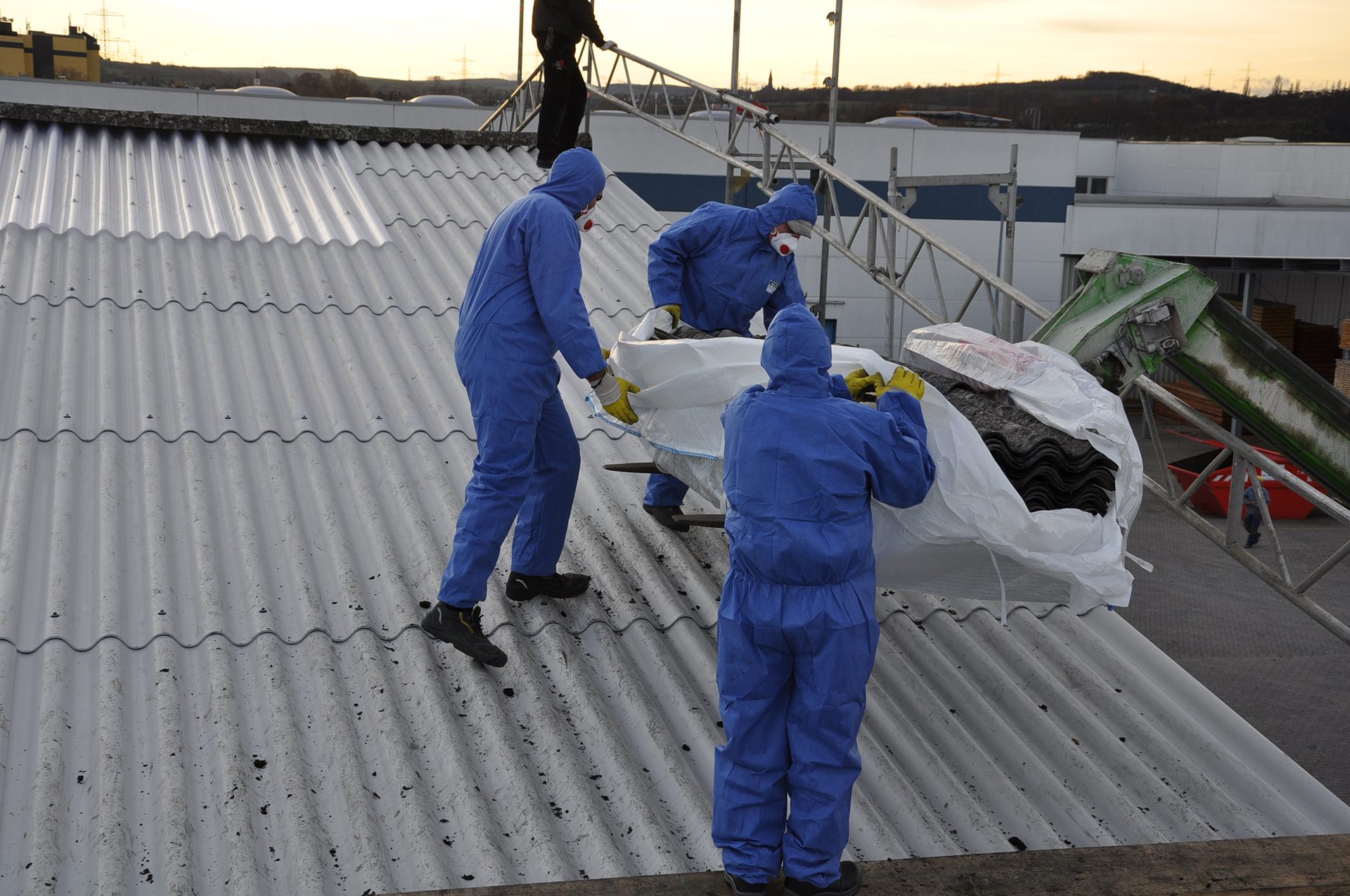 Ilustracja przedstawia mężczyzn w ochronnych kombinezonach zdejmujących azbest z dachu.