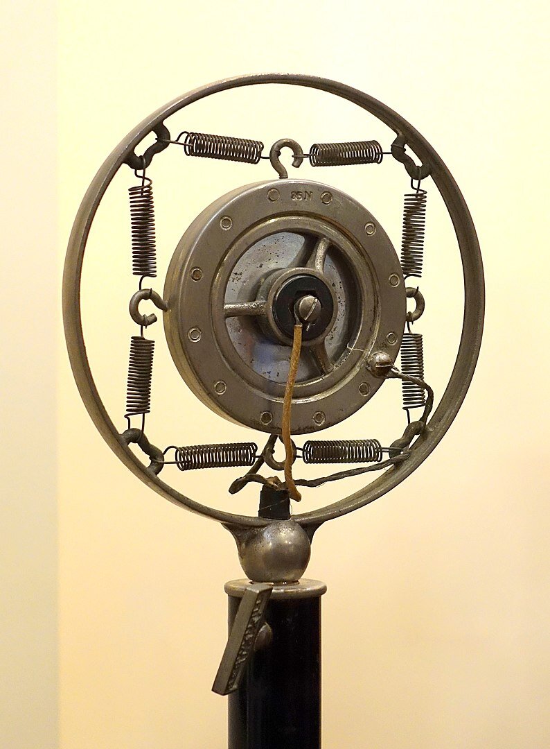 Zdjęcie przedstawia mikrofon węglowy. Na pręcie zamontowano okrąg, oraz w jego wnętrzy mniejszy. Obydwa okręgi połączono niewielkimi sprężynami z haczykami. Z okręgu środkowego widoczny jest odchodzący przewód, który przechodzi przez pręt.