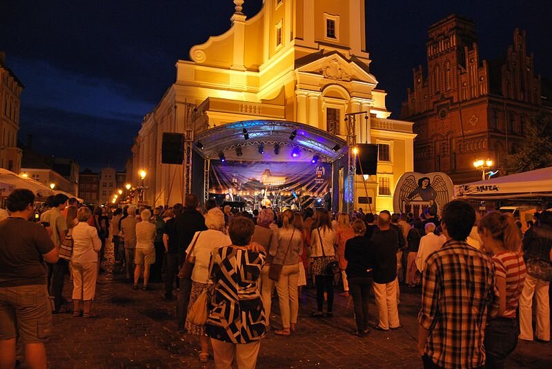 Zdjęcie przedstawia toruński rynek. Jest wieczór, przed kościołem stoi scena, na której odbywa się koncert. Przyglądają się mu licznie zgromadzeni na rynku ludzie.