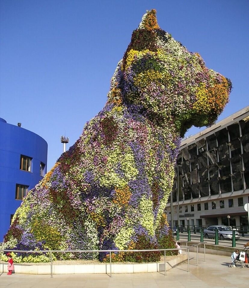 Fotografia przedstawia rzeźbę - psa, na tle niebieskiego budynku stojącego z lewej strony, jasnego budynku z oszklonym frontem z prawej strony, a także bezchmurnego, niebieskiego nieba. Rzeźba jest wielkości około 3 pięter budynku. Pies stoi w ogrodzonym fragmencie jasnego placu, na którym stoi dwójka małych dzieci. Rzeźba została wykonana na stelażu ze stali nierdzewnej, na której umieszczono doniczki z kwiatami. Przeważają kolory żółty, zielony oraz fioletowy, przypominające łaty na umaszczeniu zwierzęcia. Pies jest w pozycji siedzącej, z niewielkimi uszami oraz masywnym pyskiem. Rzeźba psa została uchwycona na fotografii z boku.