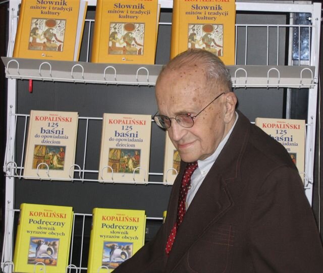 Kolorowa fotografia przedstawia starszego, łysego mężczyznę w okularach, ubranego w brązową marynarkę, białą koszulę i czerwony krawat w białe grochy. Pozuje do zdjęcia na tle napisanych przez siebie słowników i innych książek: Słownik mitów i tradycji kultury, Podręczny słownik wyrazów obcych, 125 baśni do opowiadania dzieciom.