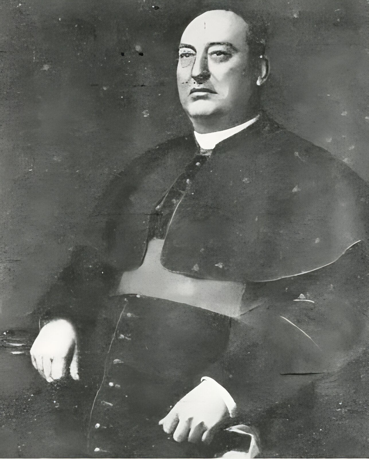 Zdjęcie przedstawia siedzącego w fotelu tęgiego mężczyznę w sutannie. Szata liturgiczna wraz z pelerynką narzuconą na ramiona, wskazuje, że pełnił on wysokie godności w kościele katolickim. Mężczyzna ma krótko obcięte włosy i okrągłą, pucołowatą twarz. 