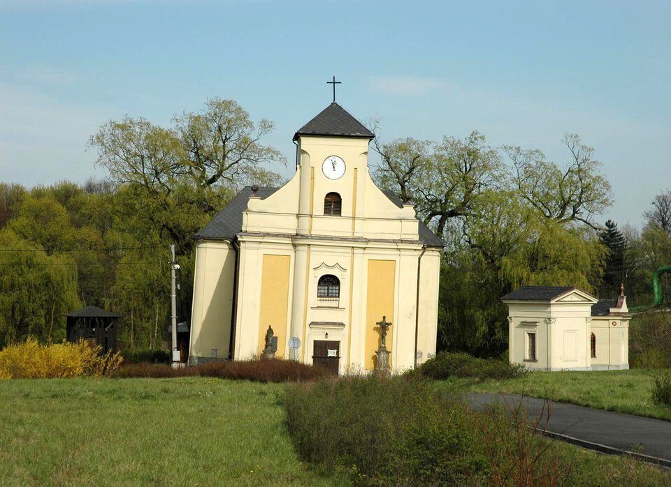 Na zdjęciu jest kościół stojący w terenie zielonym. Budynek kościoła jest nieznacznie odchylony od pionu w prawą stronę. Kościół jest niewysoki, zwieńczony wieżą z krzyżem, na wieży jest zegar. Fasada kościoła pomalowana jest na żółto. Dookoła obiektu rośnie trawa i niskie krzewy. Do kościoła prowadzi chodnik. W tle rosną drzewa.  