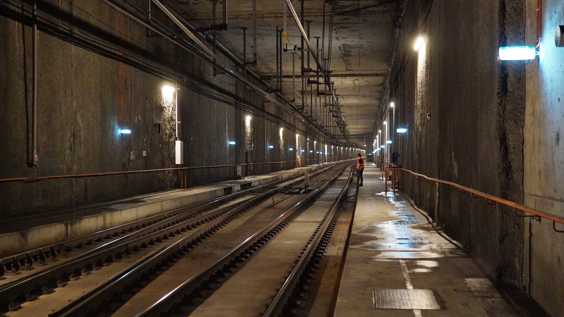 Zdjęcie przedstawia Tunel kolejowy. Wykonany jest z betonu, a jego wnętrze jest dobrze oświetlone, urządzenia oświetlające zamontowane są na ścianach tunelu. Widać dwa tory oraz w tle ludzi w odblaskowych kamizelkach, którzy pracują na linii kolejowej. Obok torowiska znajdują się chodniki, z których korzystają pracownicy kolei. Na suficie tunelu widoczne są wiszące metalowe elementy i linie zasilające.