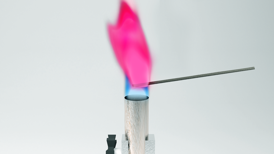 Ilustracja przedstawia metalowy pręt nad płomieniem palnika. Płomień ma kolor karminowy, czyli intensywnie różowo‑czerwony. 