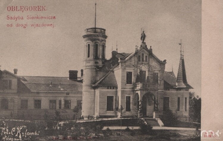 Fotografia przedstawia pałacyk. Budowla ma trzy kondygnacje. Wejście znajduje się pośrodku budynku, jest ono zadaszone. Zadaszenie jest oparte na dwóch kolumnach. Wiedzie do niego kilka schodów. Nad wejściem umieszczona jest rzeźba wyobrażająca postać w obszernej szacie. Po prawej stronie zdjęcia znajduje się czworokątny wykusz zwieńczony wieżą. W głębi, po lewej stronie stoi okrągła wieża z wąskimi oknami. Na jej umieszczony jest dość wysoki maszt.
