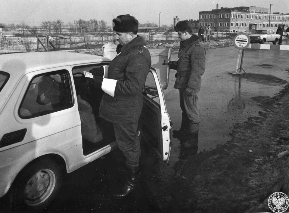 Zdjęcie przedstawia ulicę. Stoi niej samochód marki Maluch. Przy otwartych drzwiach od strony pasażera stoi milicjant, który przegląda dokumenty. Drugi milicjant zagląda do bagażnika samochodu. Kilka metrów przed samochodem stoi znak z napisem Kontrola drogowa MO. W tle znajduje się inny samochód osobowy, rozległe pole i duży budynek.