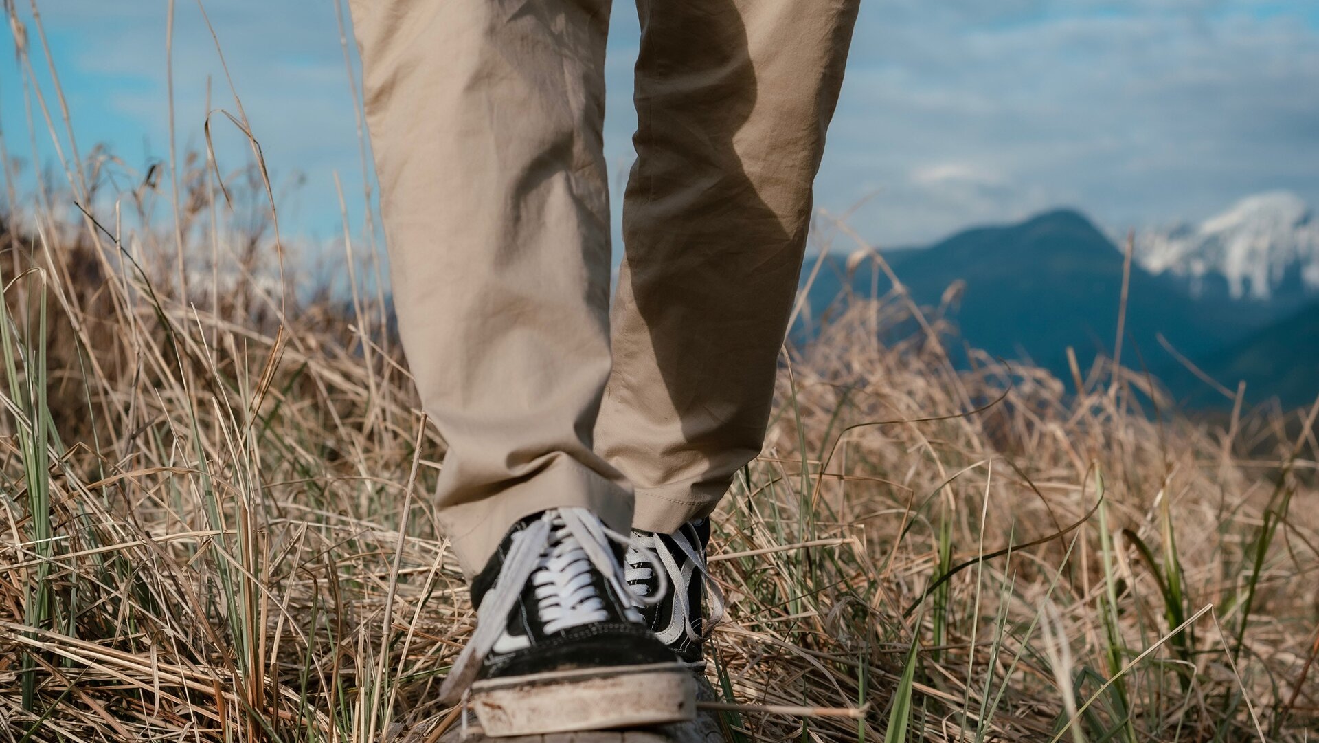Zdjęcie przedstawia nogi idącej osoby, w ujęciu od stóp do kolan. Na nogach widoczne są czarne trampki i beżowe długie spodnie. Postać idzie lub stoi na wysuszonej trawie. W tle po prawej widoczne są szczyty, w tym jeden ośnieżony. 