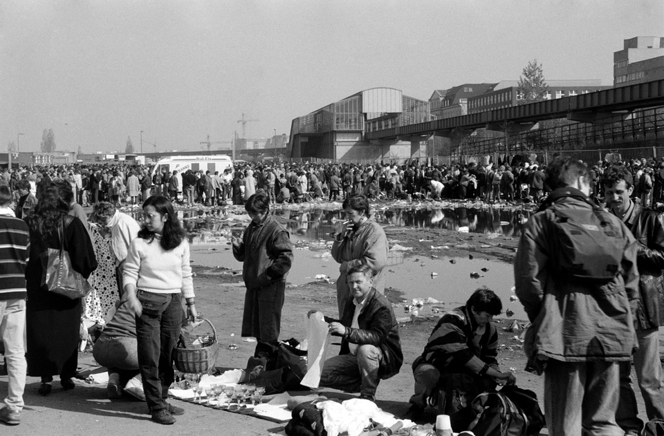 Zdjęcie przedstawia dużo ludzi na obszernym placu. Na ziemi na kocach, kawałkach materiałów leżą różne rzeczy do sprzedania. W środkowej części placu jest duża kałuża i leży dużo śmieci.    