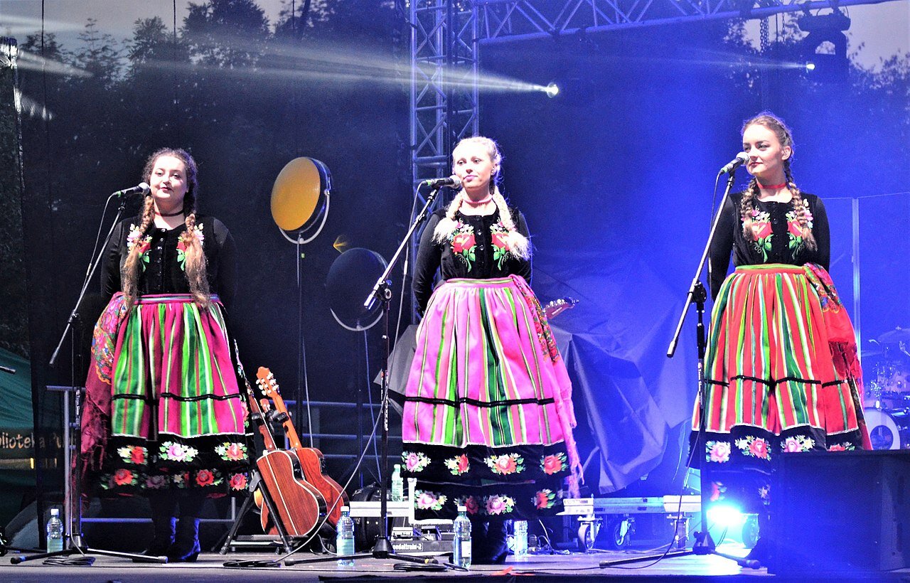 Zdjęcie przedstawia trzy artystki z zespołu Tulia. Młode kobiety występują na scenie. Są ubrane w stroje ludowe.