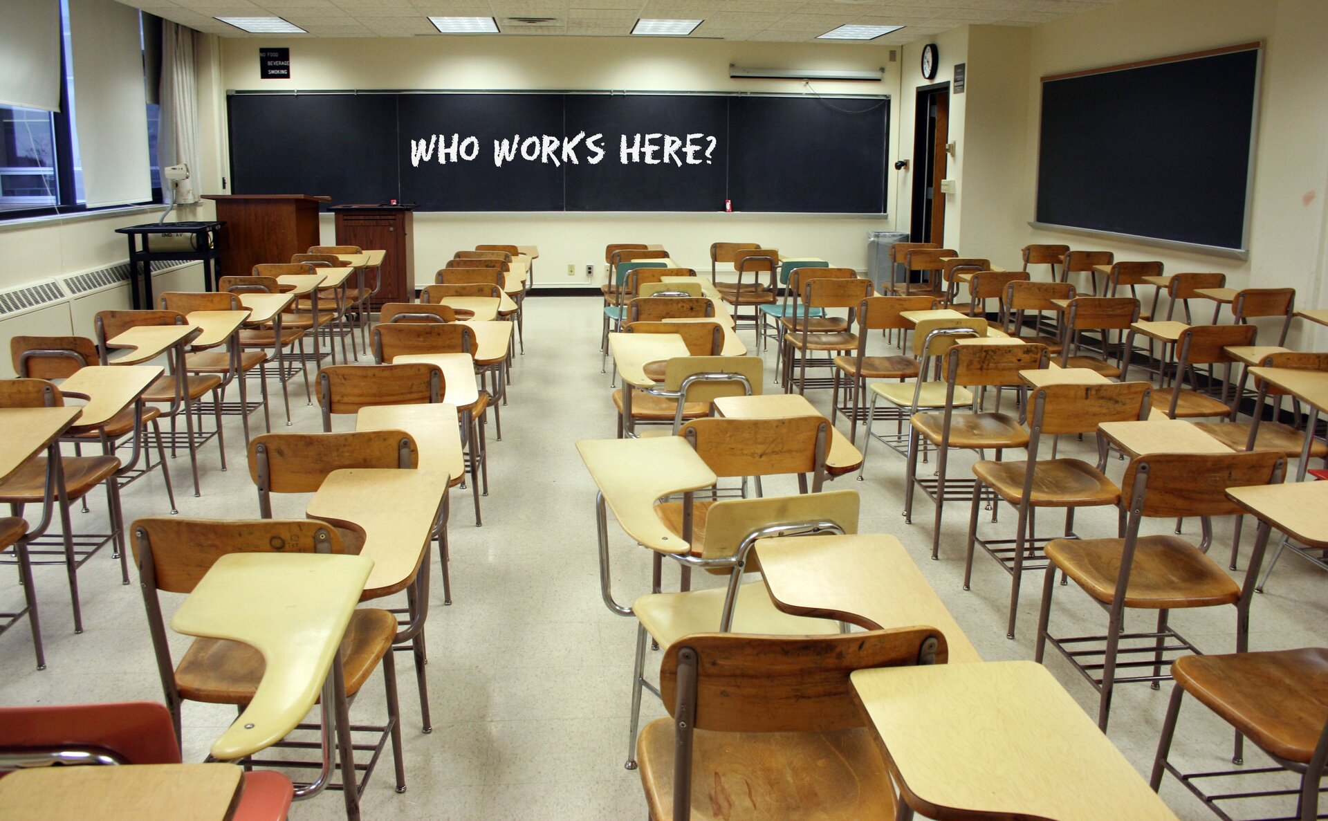 Zdjęcie przedstawia pustą salę lekcyjną, wypełnioną szkolnymi ławkami z krzesłami. Na czarnej kredowej tablicy jest biały napis "WHO WORK'S HERE?"