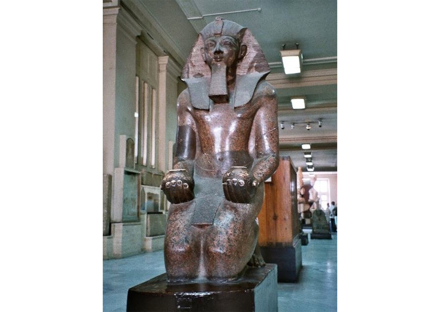 Na zdjęciu statua Faraona z granitu w muzeum. Mężczyzna siedzi. Dłonie ma oparte na udach. W każdej dłoni trzyma małe, okrągłe naczynie.. 