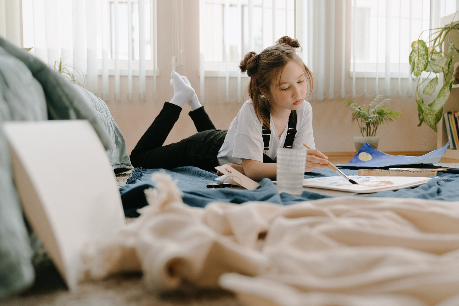 Ilustracja przedstawia dziewczynkę, która leży na podłodze i maluje farbami obrazek.