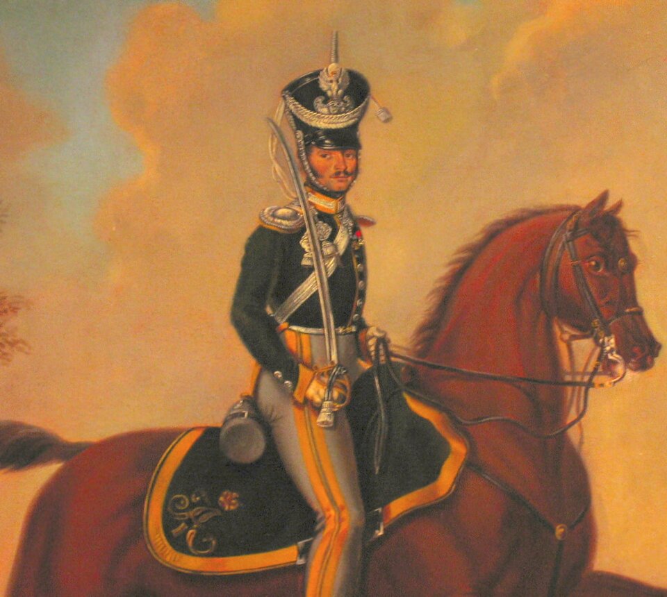 Ilustracja przedstawia młodego mężczyznę na koniu. Ubrany jest w mundur: krótką kurtkę ze srebrnymi guzikami, białożółtym kołnierzem typu stójka i srebrnymi, okrągłymi naramiennikami. Przez lewe ramię ma przerzuconą srebrną szarfę, a ponad nią na lewej piersi ma odznaczenie wojskowe. Ma szare spodnie z żółtymi lampasami. Na dłoniach ma białe rękawiczki. W prawej ręce trzyma uniesioną do góry szablę. Na głowie ma wysoką, usztywnioną czapkę z okrągłym denkiem i daszkiem przyozdobioną srebrnym sznurem i orłem na przodzie. Do czapki przytwierdzony jest srebrny sznur zakładany pod brodę. Pod siodłem jeźdźca umieszczona jest zielona, ozdobna derka z żółtym lampasem. 