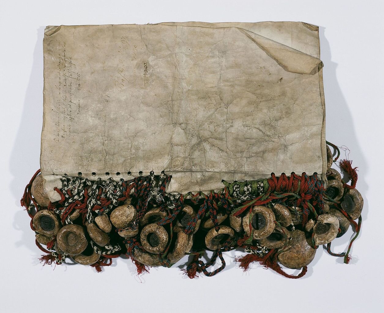 Kliknij, aby powiększyć. Zdjęcie przedstawia pożółkłe kartki papieru ułożone na białym tle. Papier jest częściowo zapisany czarną, ozdobną czcionką. Napisy są nieczytelne. Kartki leżą w poziomie. Dół kartek został podziurkowany, a w każdej z dziur znajduje się czerwony, biało‑czarny lub brązowy sznur, do którego przypięte są pieczęcie. Są one okrągłe z pogrubionymi bokami. Są pożółkłe, a ich środek jest czarny.