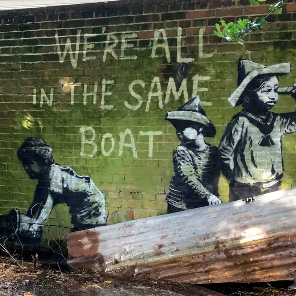 Fotografia przedstawia pracę Banksy’ego znajdującą się na ścianie w Nicholas Everitt Park. Na zielonej ścianie techniką szablonu namalowano trzech młodych chłopców, ubranych o marynarsku, znajdujących się na łodzi. Dzieci mają na głowach szpiczaste kapelusze. Najwyższy chłopiec stoi po prawej stronie. W lewej ręce trzyma lunetę, przez którą patrzy w dal. Prawą rękę trzyma za plecami. Tuż za nim stoi niższy chłopiec, który głowę odwróconą ma za siebie i patrzy na trzeciego chłopca, który wiaderkiem wybiera wodę z łodzi. Nad chłopcami umieszczono napis „We’re all in the same boat”, czyli „Wszyscy jesteśmy na tej samej łodzi”. Przy ścianie umieszczono fragment brązowej, falistej blachy, przypominającej burtę łodzi.