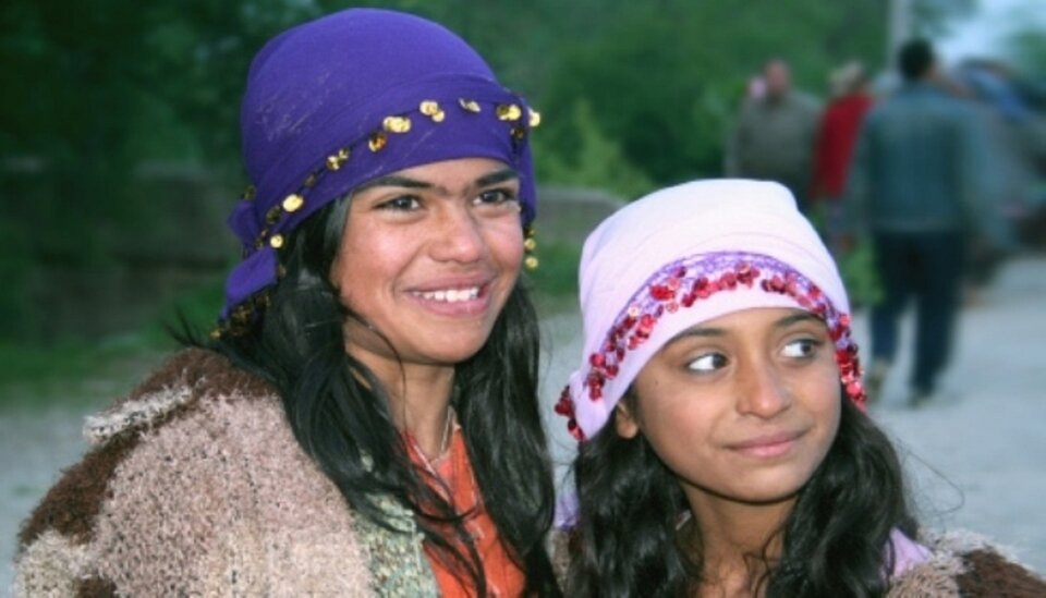 Ilustracja przedstawia dwie romskie dziewczyny. Mają na sobie ozdobne okrycia głowy i szaro‑brązowe swetry.