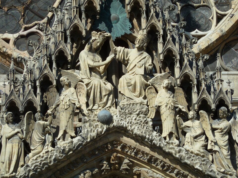 Ilustracja przedstawia rzeźby na przyczółku zachodniego portalu katedry w Reims. Ukazuje scenę koronacji Marii, zamieszczoną w wimperdze nad archiwoltami. Pośrodku siedzi Maria i Chrystus w koronie, nakładający na Jej głowę koronę. Po bokach scenę adorują aniołowie. Nad rzeźbami portalowymi znajdują się gotyckie, ostro zamknięte baldachimy w postaci arkadek, wspartych na cienkich kolumienkach.  