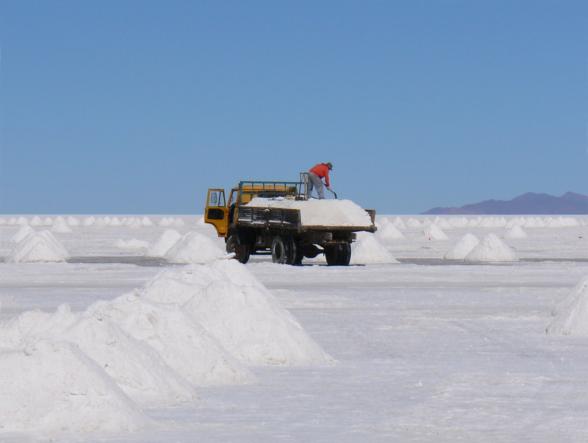 Zdjęcie przedstawia solnisko Salar de Uyuni w Boliwii. Na płaskiej, białej płaszczyźnie usypane są pryzmy soli. W centrum znajduje się żółta wywrotka z przyczepą załadowaną solą. Na przyczepie stoi człowiek z łopatą w ręce.