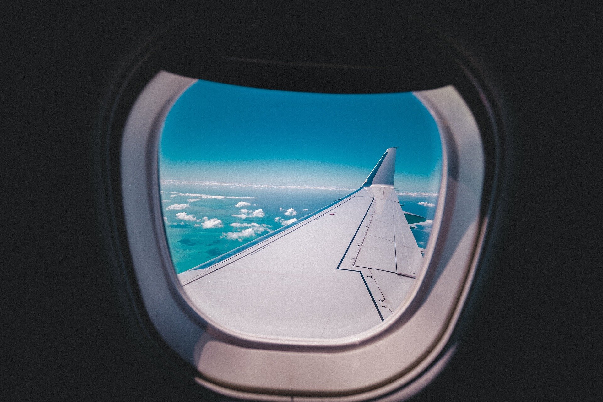 Zdjęcie przedstawiające okno samolotu z widokiem na skrzydło błękitne niebo, poniżej samolotu zaś oświetlone słońcem chmury.