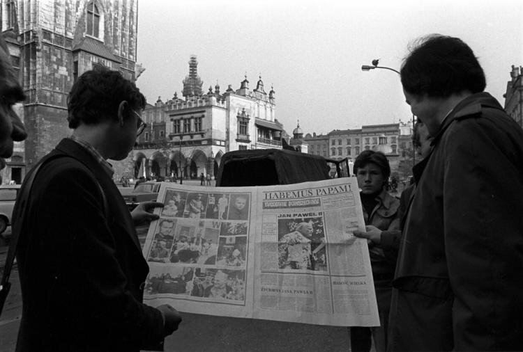 Zdjęcie przedstawia krakowski Rynek. Stoją na nim dwaj mężczyźni trzymający rozłożoną gazetę. Widnieje na niej duży nagłówek Habemus Papam!.