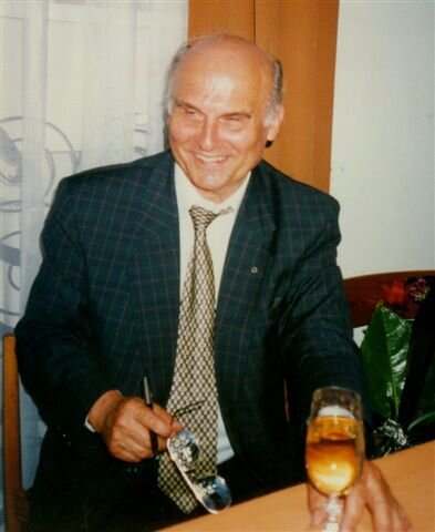 Zdjęcie przedstawia siedzącego przy stole mężczyznę w średnim wieku. Nosi elegancki ciemny garnitur, białą koszulę i srebrny krawat. Ma okrągłą, opaloną, uśmiechniętą twarz. Siwe, krótkie włosy okalają po bokach rozległą łysinę. W lewej ręce trzyma okulary i wieczne pióro. Palcami prawej dłoni dotyka kieliszka z winem. W tle znajduje się okno z firanką i zasłoną.