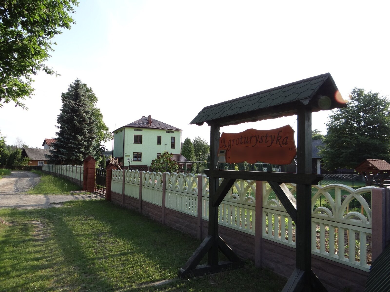 Na fotografii zielony, piętrowy budynek mieszkalny, otoczony murowanym ogrodzeniem. Przed ogrodzeniem szyld z drewnianą tabliczką. Na niej napis: Agroturystyka Pod Lasem. Wokół trawa, w oddali inne domy.