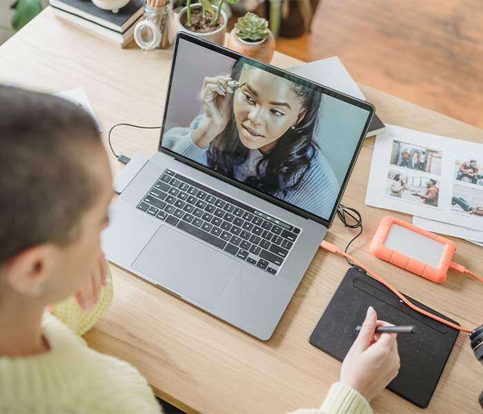 Zdjęcie pierwsze przedstawia wykonywanie retuszu fotografii. Widoczny jest na nim mężczyzna siedzący przy biurku, na którym stoi otwarty laptop. Na laptopie włączone jest zdjęcie przedstawiające kobietę. Na biurku obok laptopa leży urządzenia podpięte do komputera za pomocą przewodu.