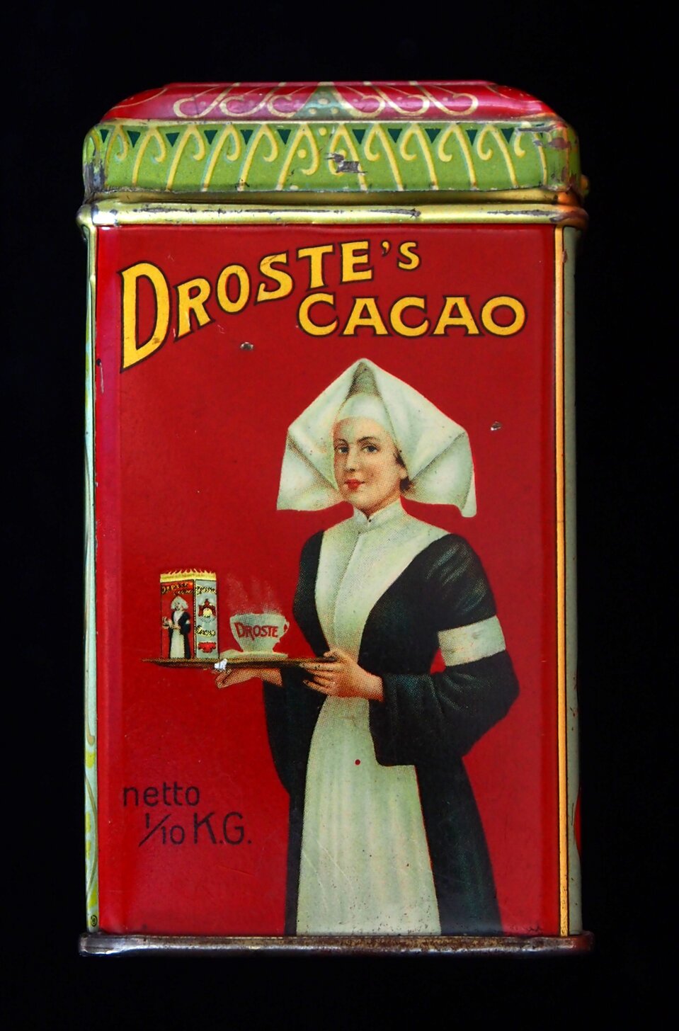 Fotografia przedstawia puszkę z kakao firmy:  Droste's cacao .  Znajduje się na nim kobieta trzymająca to samo opakowanie kakao oraz filiżankę na spodku z napisem:  Droste . Kobieta ubrana jest w biały czepek, długą suknię zapiętą pod szyję z pionowym pasem biegnącym przez środek od kołnierzyka w dół i białą opaską na lewym rękawie. W lewym dolnym rogu opakowania znajduje się napis: netto 1/10 K.G., Tło posiada kolor czerwony. 