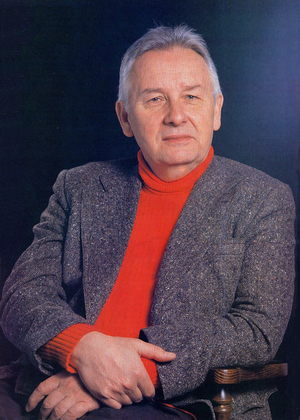 Fotografia przedstawia: Henryk Mikołaj Górecki, siedzący na krześle. Kompozytor ma ciemną marynarkę oraz czerwony sweter z golfem i siwe, krótkie włosy.