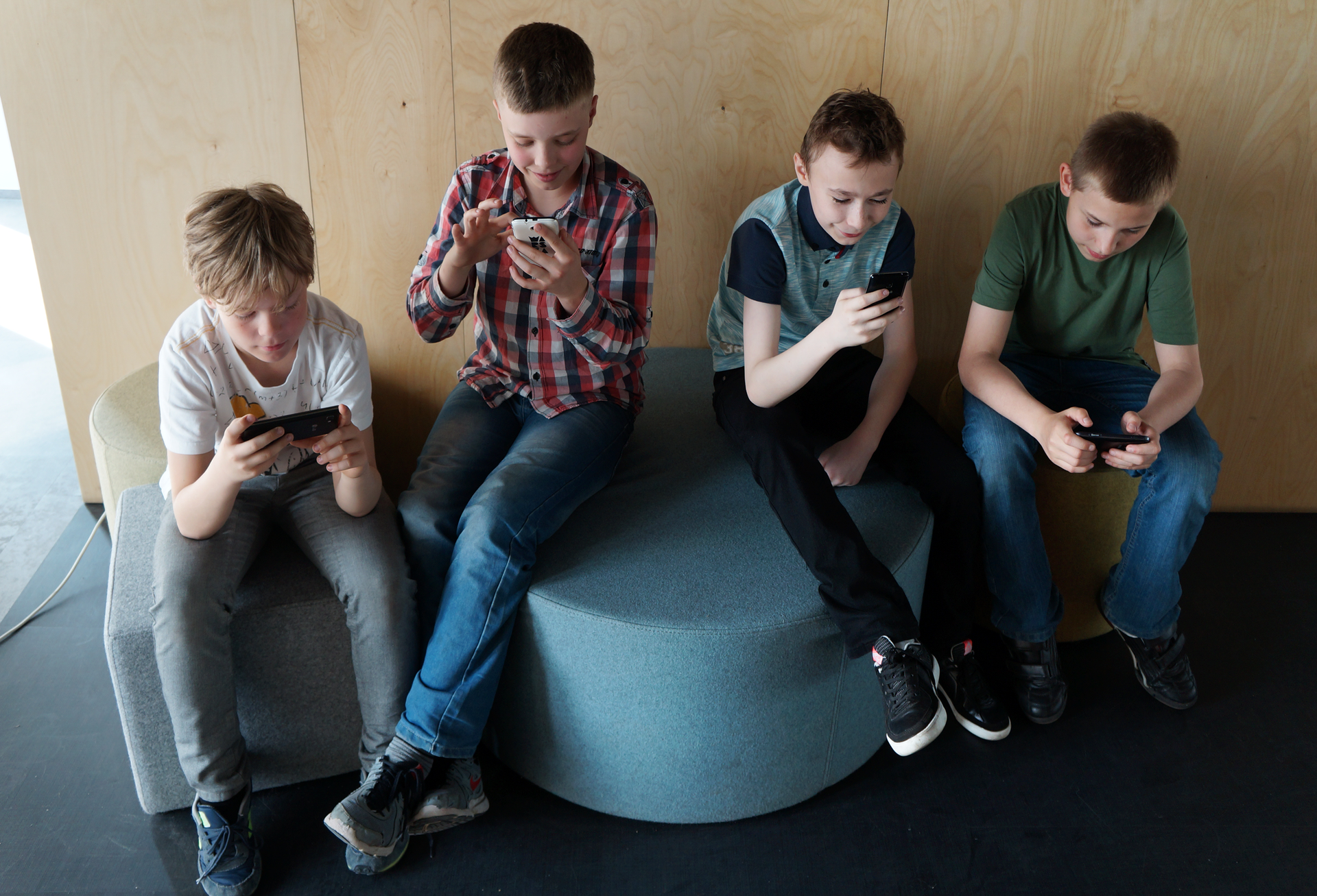 Fotografia przestawia widzianą z góry grupę czterech chłopców w różnokolorowych strojach. Mają około 10‑12 lat. Siedzą na pufach. Każdy z nich patrzy w ekran swojego telefonu, odczytuje lub wysyła wiadomości.