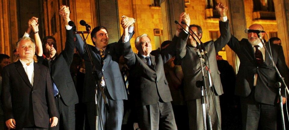 Ilustracja przestawia moment przemówienia w 2008 roku. Polski polityk stoi pierwszy od lewej. Jest ubrany w biała koszulę bez krawata, która jest rozpięta pod szyją. Ma na sobie ciemną marynarkę. Włosy są przyprószone siwizną. Jest on niższy od pozostałych postaci. Obok polskiego polityka stoją czterej mężczyźni ubrani w ciemne garnitury. Trzymają się za ręce, które są uniesione w górę w geście solidarności. 