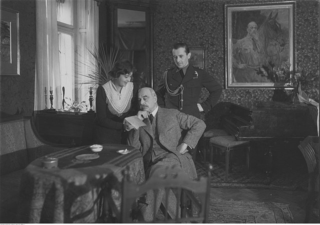 Zdęcie przedstawia wnętrze salonu. Na pierwszym planie, przy niewielkim, owalnym stole siedzi starszy mężczyzna w garniturze. Ma założoną nogę na nogę. Prawy łokieć wspiera na stole. Przy prawym boku mężczyzny stoi dojrzała kobieta w sukni. Pokazuje starszemu mężczyźnie kartkę papieru, uśmiecha się. Za plecami siedzącego mężczyzny stoi dojrzały mężczyzna w wojskowym mundurze. Trzyma podparte na bokach dłonie. W tle postaci sofa, fortepian i dwa stojące obok niego taborety. Na ścianach zawieszone są obrazy, w rogu salonu wysokie tremo.