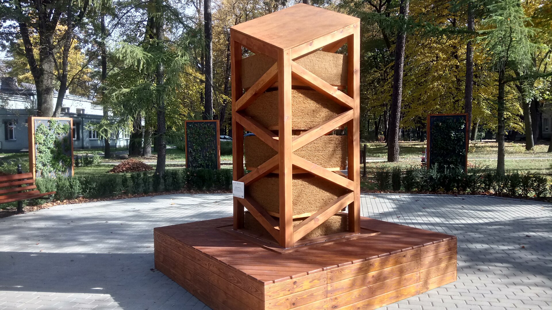 Na zdjęciu widoczna niewysoka drewniana wieża na platformie, stojąca w parku. Wieża ma ażurowe boki, formują je belki ułożone skośnie. W środku widoczne bloki z włókna kokosowego.