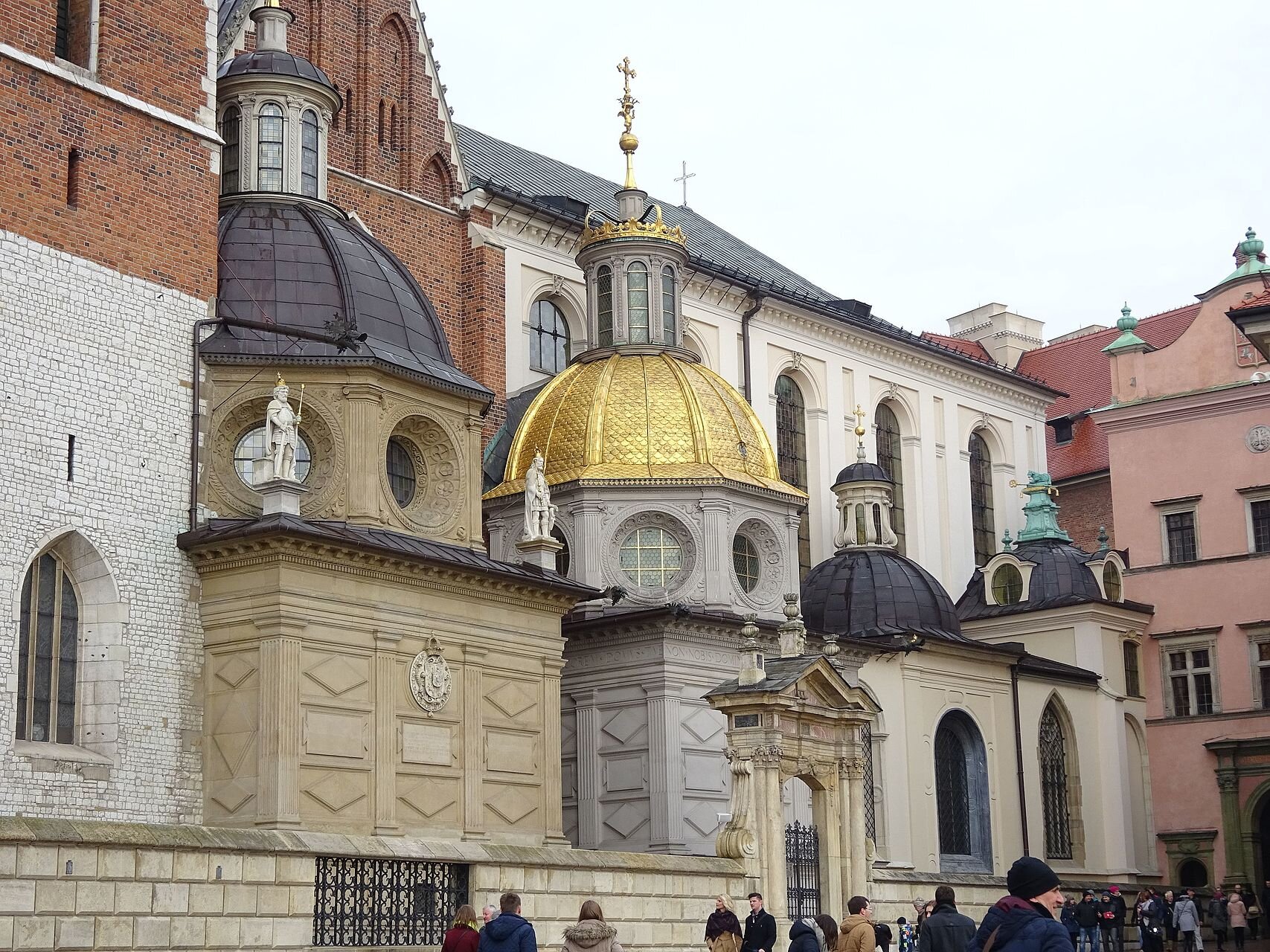 Na zdjęciu znajdują się kaplice katedry wawelskiej. Są one dobudowane do ściany katedry. Są to wysokie budynki z czarnymi i lub złotymi kopułami. Przed wejściem do nich znajduje się grupa zwiedzających. 