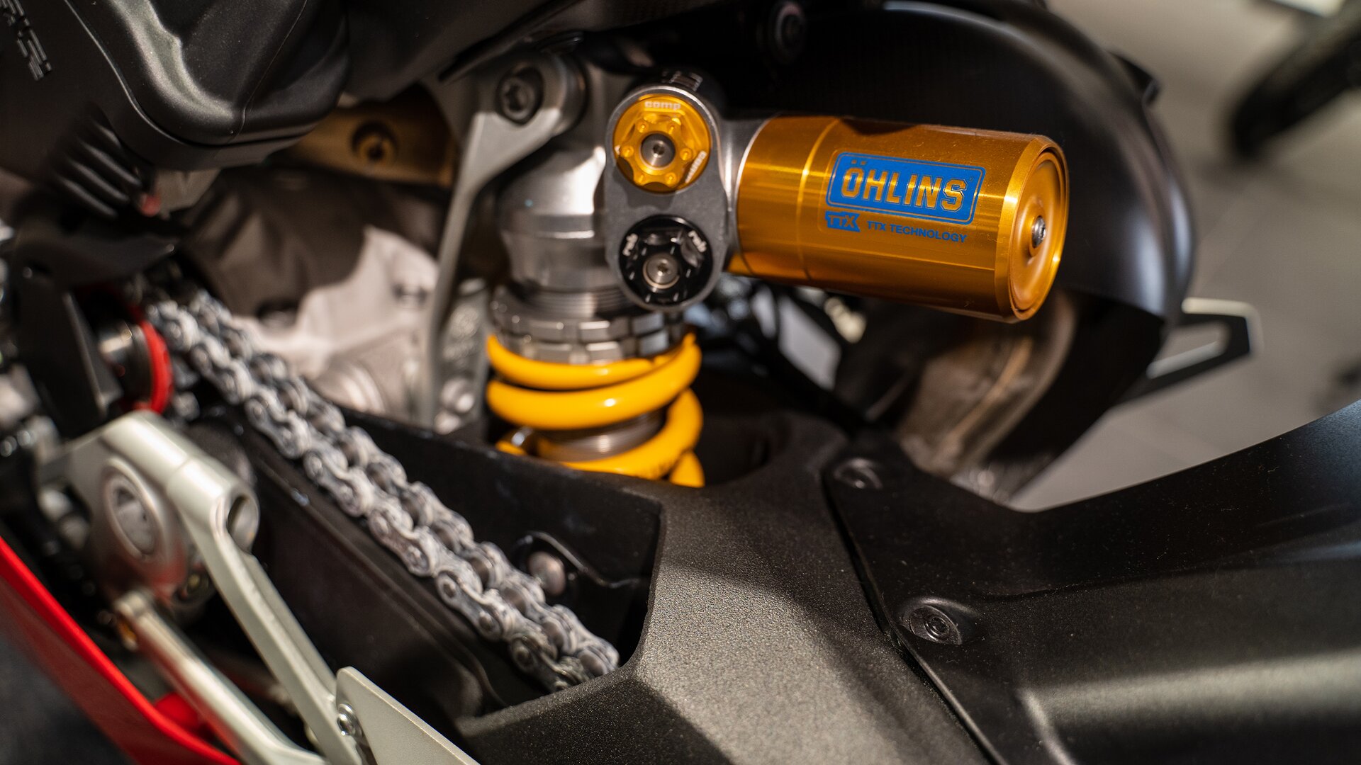 Zdjęcie pokazuje zbliżenie na element w kształcie metalowego wałeczka. Na nim napis OHLINS. Jest połączony pod kątem dziewięćdziesięciu stopni z częścią, wokół której jest żółta gruba sprężyna.  