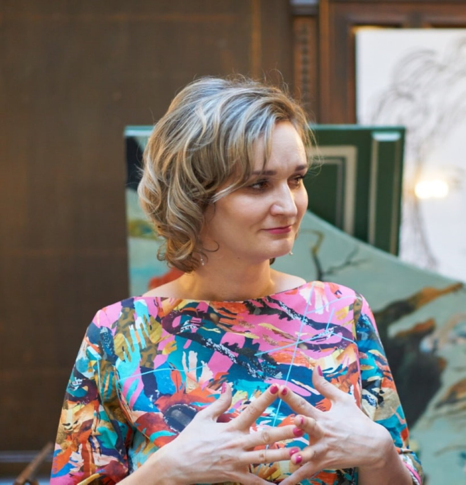 Fotografia przedstawia kobietę w średnim wieku o średniej długości blond włosach, która została uchwycona z profilu. Kobieta spogląda się w prawą stronę. Ubrana jest w kolorową sukienkę o abstrakcyjnym wzorze - składającym się z plam barwnych. Dłonie ma przy piersiach. 