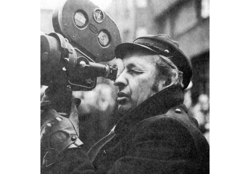 Czarno-biała fotografia przedstawiająca Andrzeja Wajdę. Ma na głowie czapkę, na dłoniach skórzane rękawiczki. W rękach trzyma dużą kamerę, przykłada do niej oko.