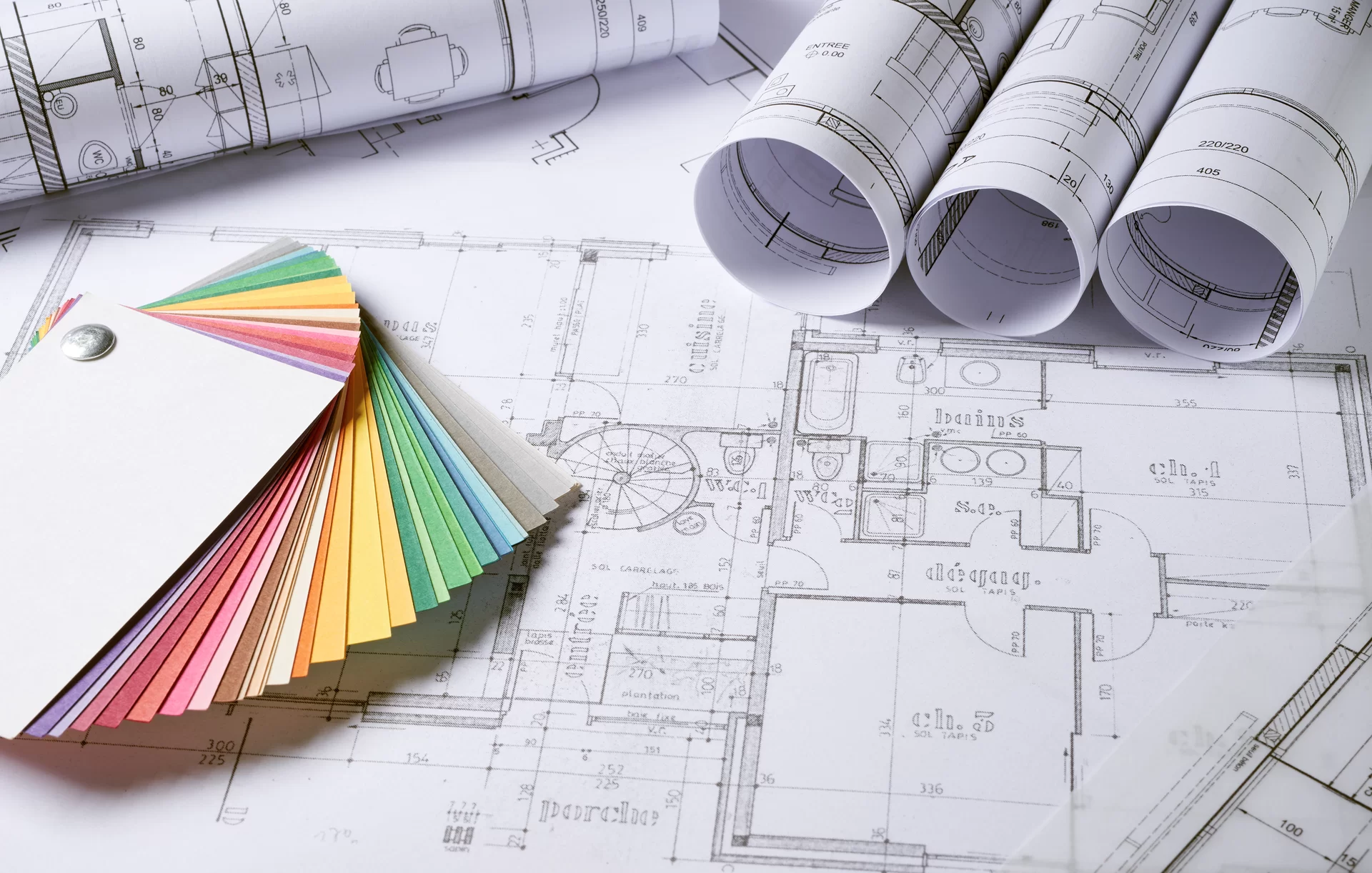 Zdjęcie przedstawia rozłożony plan architektoniczny. Po prawej stronie znajdują się cztery zwinięte plany. Po lewej stronie znajduje się próbnik kolorów składający się z wielu kolorowych prostokątnych karteczek.